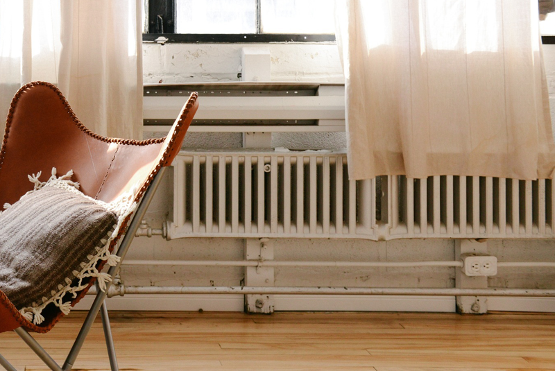 Salón antiguo con un viejo sillón en el centro y radiadores obsoletos en la pared bajo ventanas de madera. 