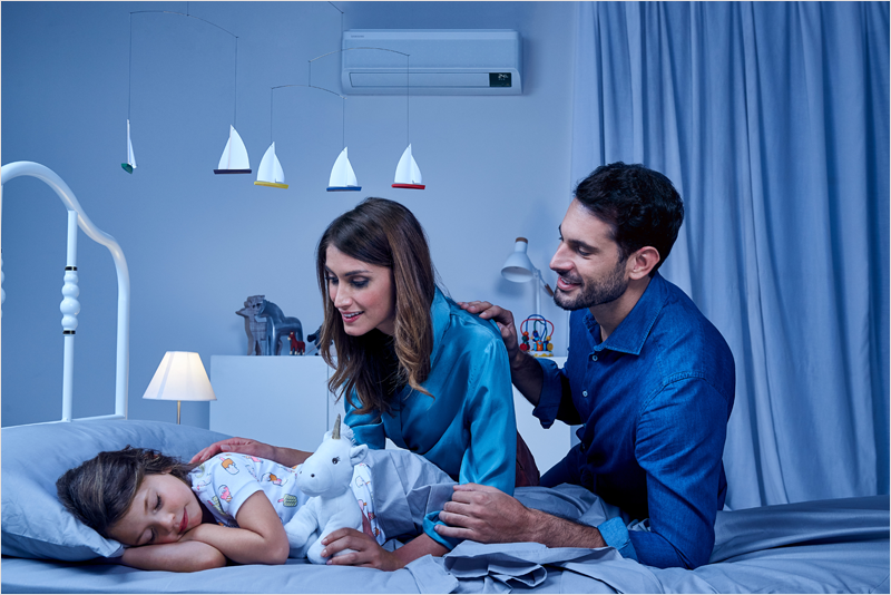 Madre y padre acostando a su hija y con un aparato de climatización de Samsung en la pared.