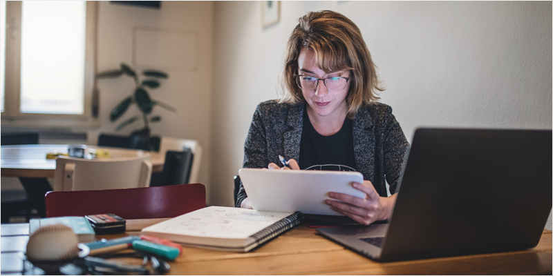 Mujer con un cuaderno y un boli mirando lo que tiene escrito. Delante de ella está también su portátil. Está en una oficina o despacho.