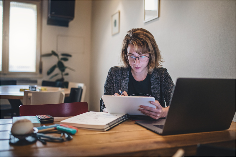 Mujer con un cuaderno y un boli mirando lo que tiene escrito. Delante de ella está también su portátil. Está en una oficina o despacho.