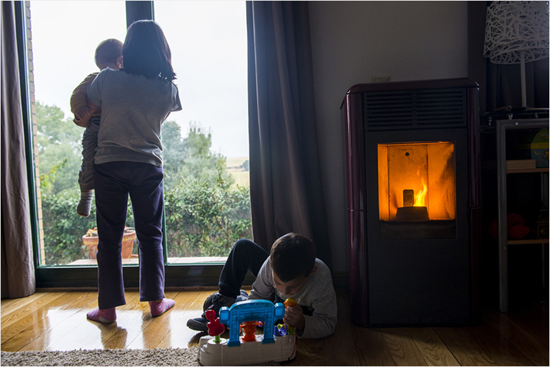 Dos niñas mirando por la ventana y un niño en el suelo jugando y en la estancia un aparato de calefacción.