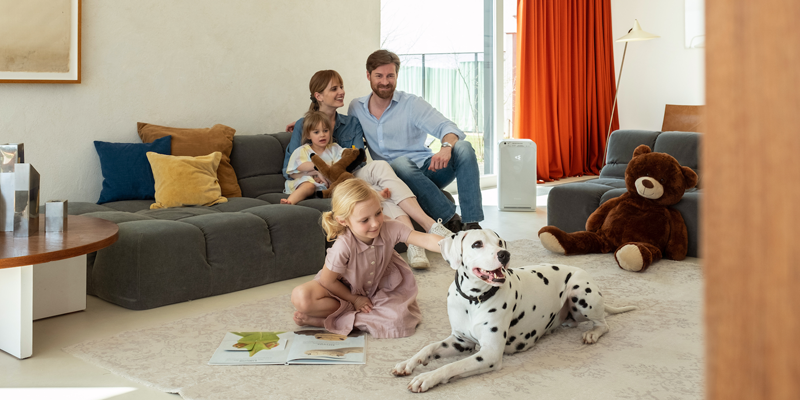 Un hombre y una mujer están sentados en el sofá con su hija pequeña mientras la mayor está en sentada en una alfombra al lado del perro y hay un aparato de ventilación de Daikin.
