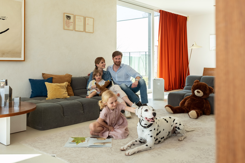 Un hombre y una mujer están sentados en el sofá con su hija pequeña mientras la mayor está en sentada en una alfombra al lado del perro y hay un aparato de ventilación de Daikin.