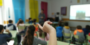 Aula con alumnos sentados en sus sillas y una chica con la mano levantada atendiendo a la profesora.