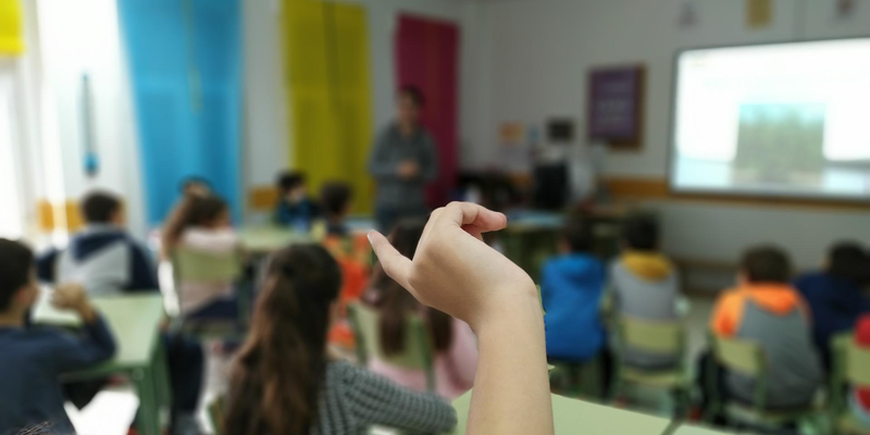Aula con alumnos sentados en sus sillas y una chica con la mano levantada atendiendo a la profesora.