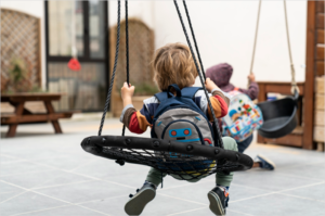 Un niño de espaldas montado un un columpio de una guardería y otro más adelante difuminado agarrando otro columpio.