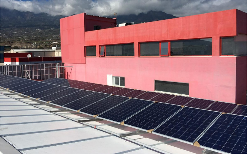 Placas solares instaladas en un tejado y al fondo un edificio rojo.
