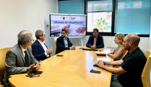 Reunión de 5 hombres y una mujer en una mesa hablando y detrás una pantalla que pone Oficinas de rehabilitación.