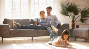 Pareja sentada en el sofá y su hija en el suelo pintando en un cuaderno.