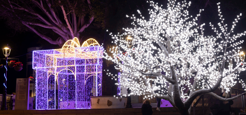 Iluminación gigante en una calle. Una caja de un regalo gigante con luces moradas y un árbol con luces blancas.