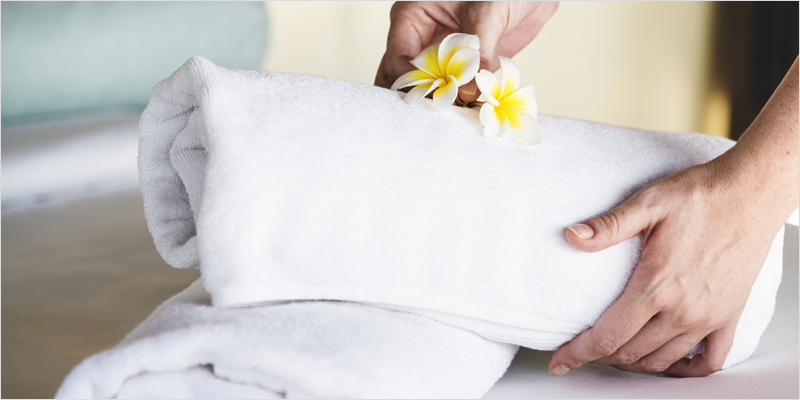 Una mano colocando dos toallas encima de una cama y una flor amarilla.