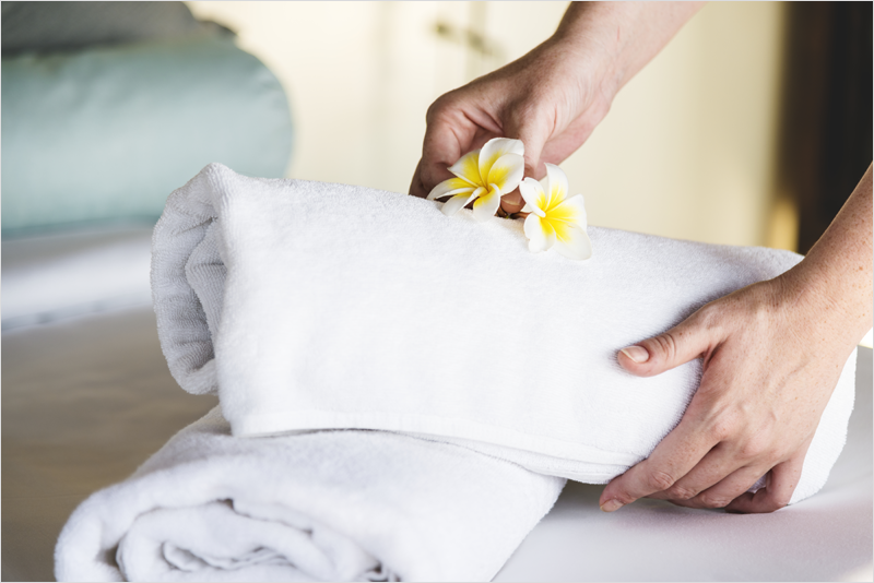 Una mano colocando dos toallas encima de una cama y una flor amarilla.