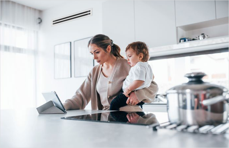 Madre con un bebé en brazos mirando una tablet en la mesa de la cocina.