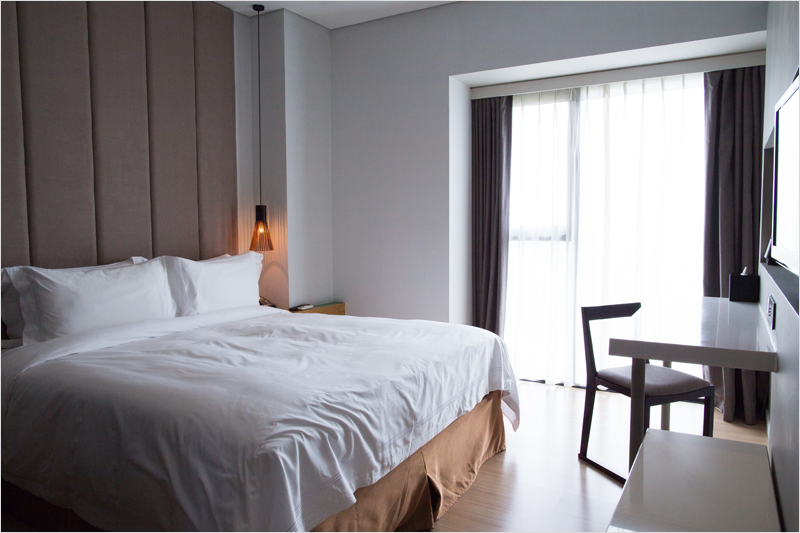 Habitación de hotel con una cama, ventana, cortinas, mesa, silla y televisión colgada en la pared.