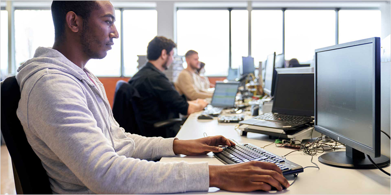 Chico en primer plano y otros dos en segundo plano sentados en sus sillas y trabajando con un ordenador.