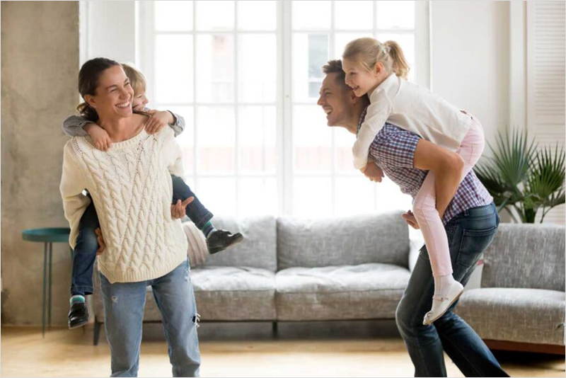 Familia en el interior de una vivienda la madre coge en la espalda al hijo y el padre a la hija. Todos sonrien.