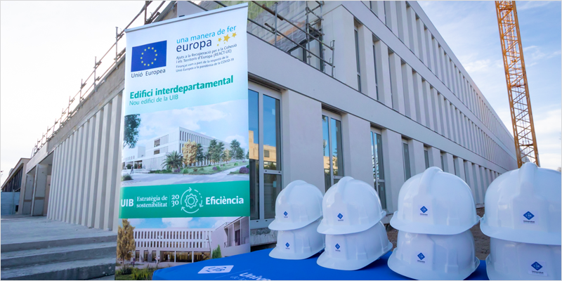 Fachada de la Universidad de Baleares y una mesa con cascos blancos y un cartel detrás que anuncia unas obras en las instalaciones.