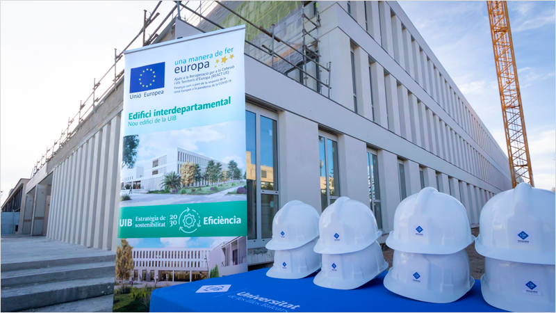 Fachada de la Universidad de Baleares y una mesa con cascos blancos y un cartel detrás que anuncia unas obras en las instalaciones.