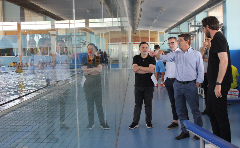 Varios hombres de pie visitando las instalaciones de una piscina cubierta.
