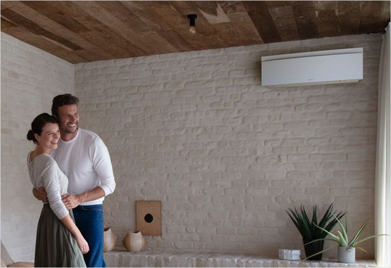 Hombre y mujer de pie abrazados en el salón de su casa y un aparato de aire acondicionado en la pared.