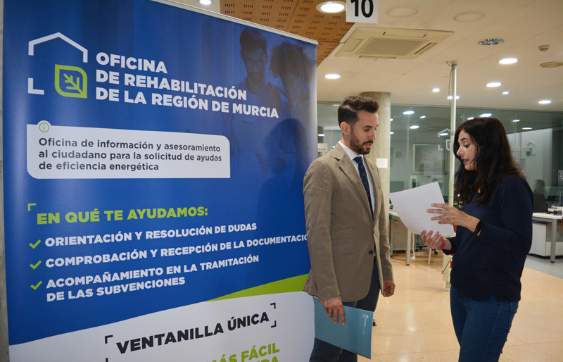 Un chico y una chica hablando de pie sobre unos papeles que tienen en la mano y detrás un cartel de la oficina de rehabilitación.