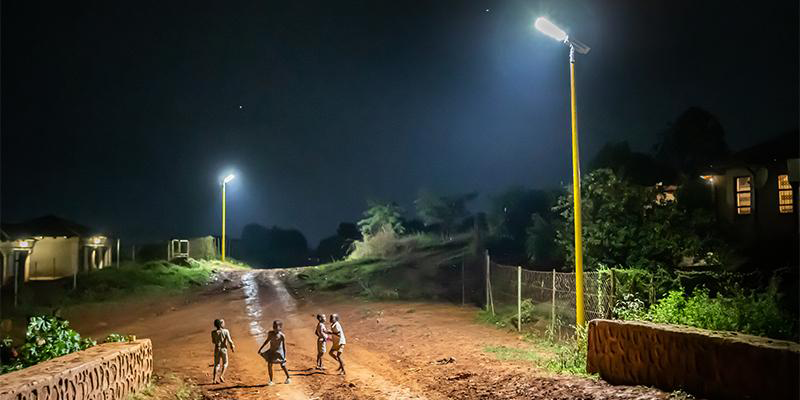 Carretera de barro y cuatro niños de color jugando en la zona que está iluminada con farolas solares con postes amarillos.