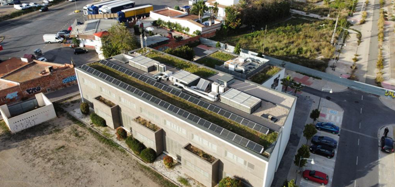Vista aérea de un edificio con placas solares en el tejado y alrededor carretera y coches aparcados.