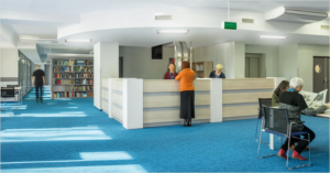 Biblioteca con libros en estanterias y personas leyendo mientras en el mostrador dos mujeres atienden a otra.