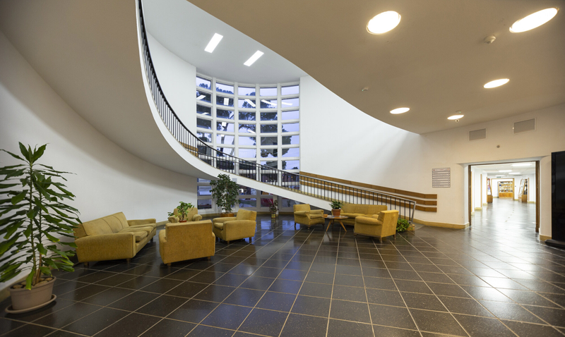 Interior de un edificio con un espacio de entrada grande y sofás, escalera y luces LED en el techo.