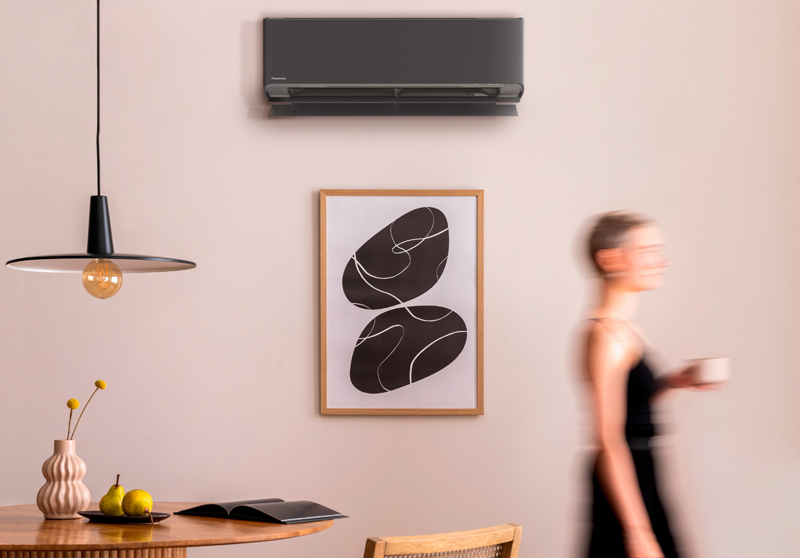 Aparato de climatización en color negro instalado en una pared y una mujer borrosa caminando con una taza de café.